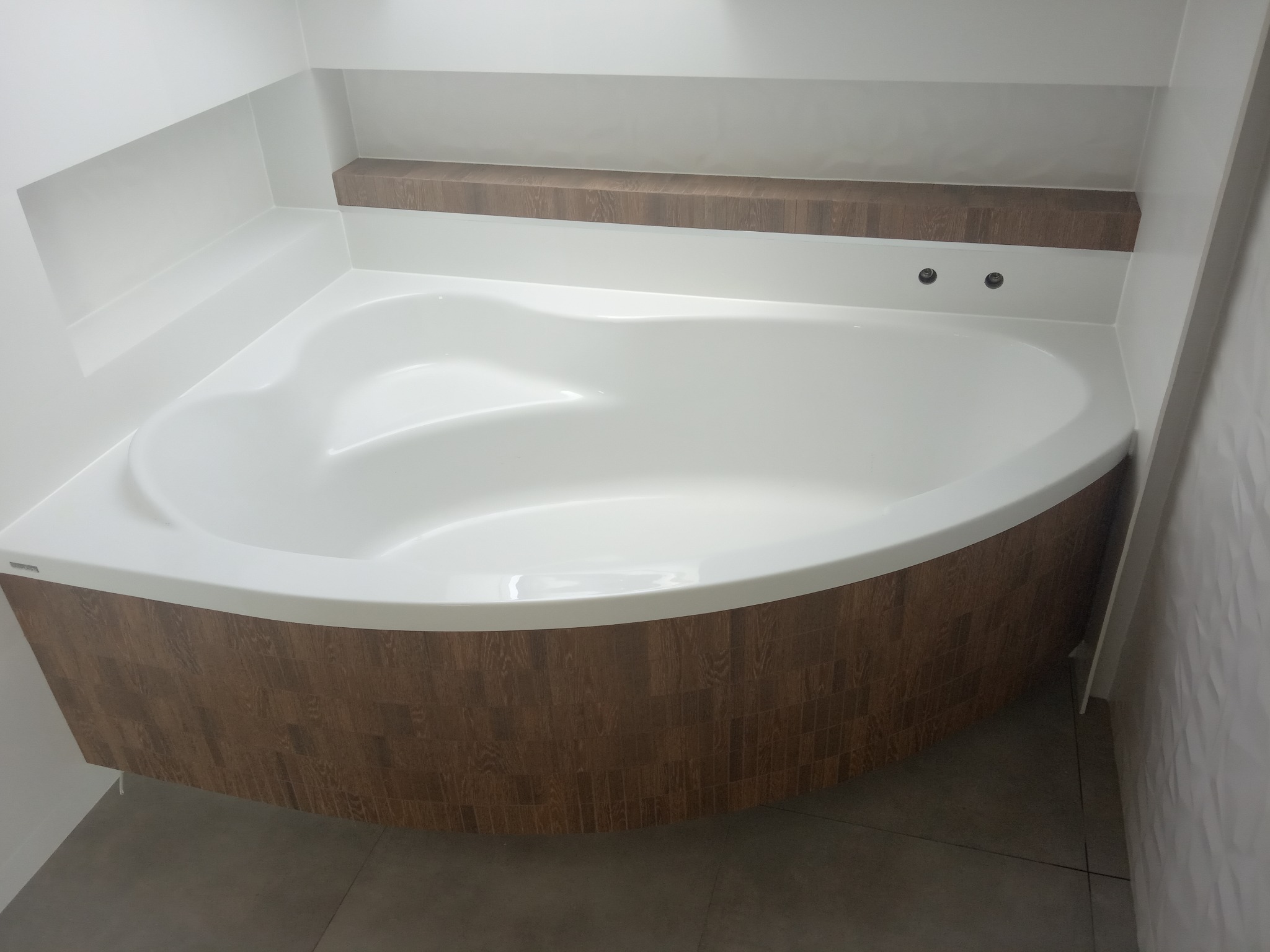 Narożna, biała wanna z obudową z paneli imitujących drewno w nowoczesnej łazience z szarymi płytkami podłogowymi i półką wnękową.