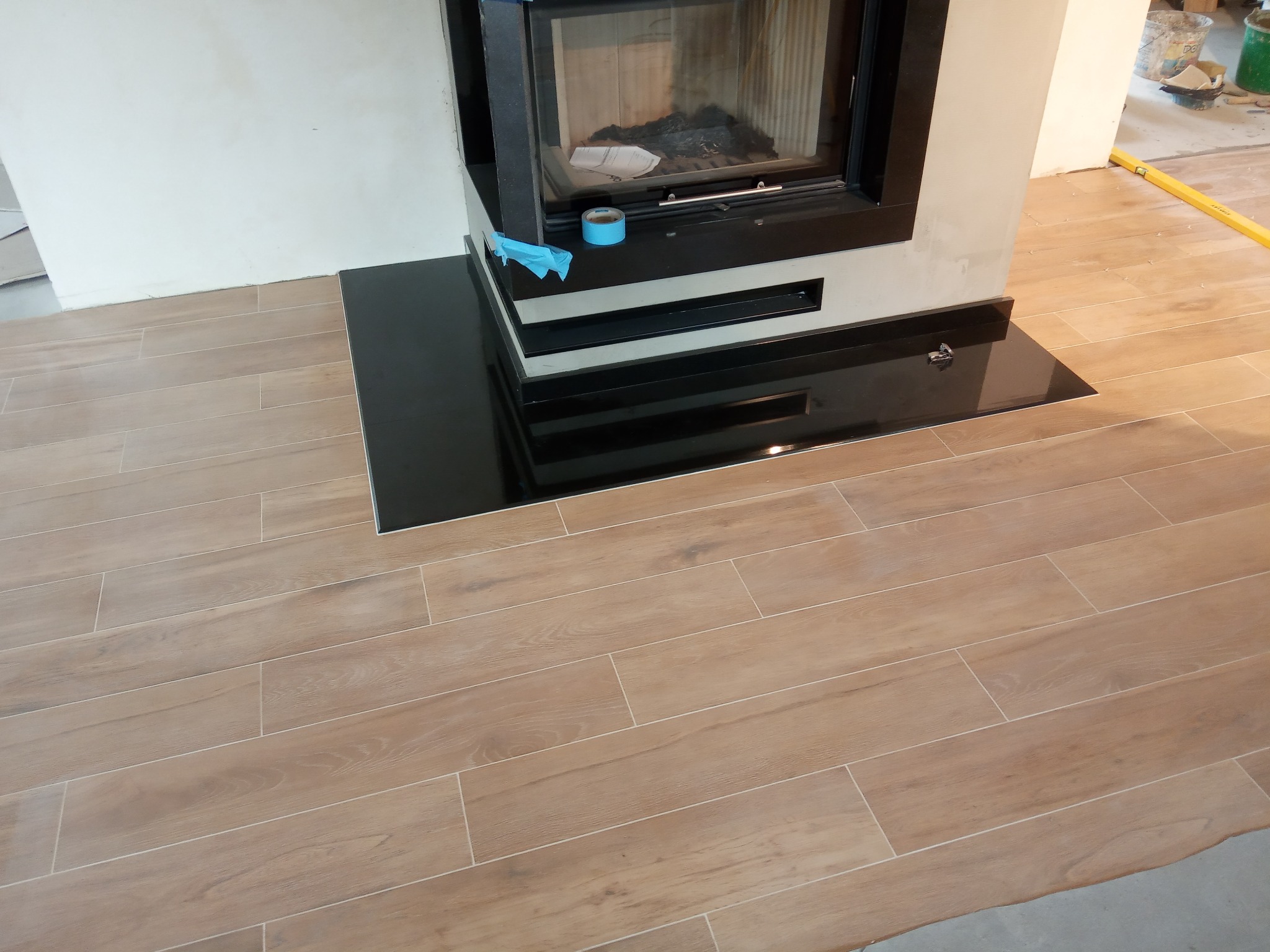 Ułożone podłużne płytki podłogowe imitujące drewno, przylegające do czarnego, błyszczącego obramowania kominka, z poziomnicą w tle.