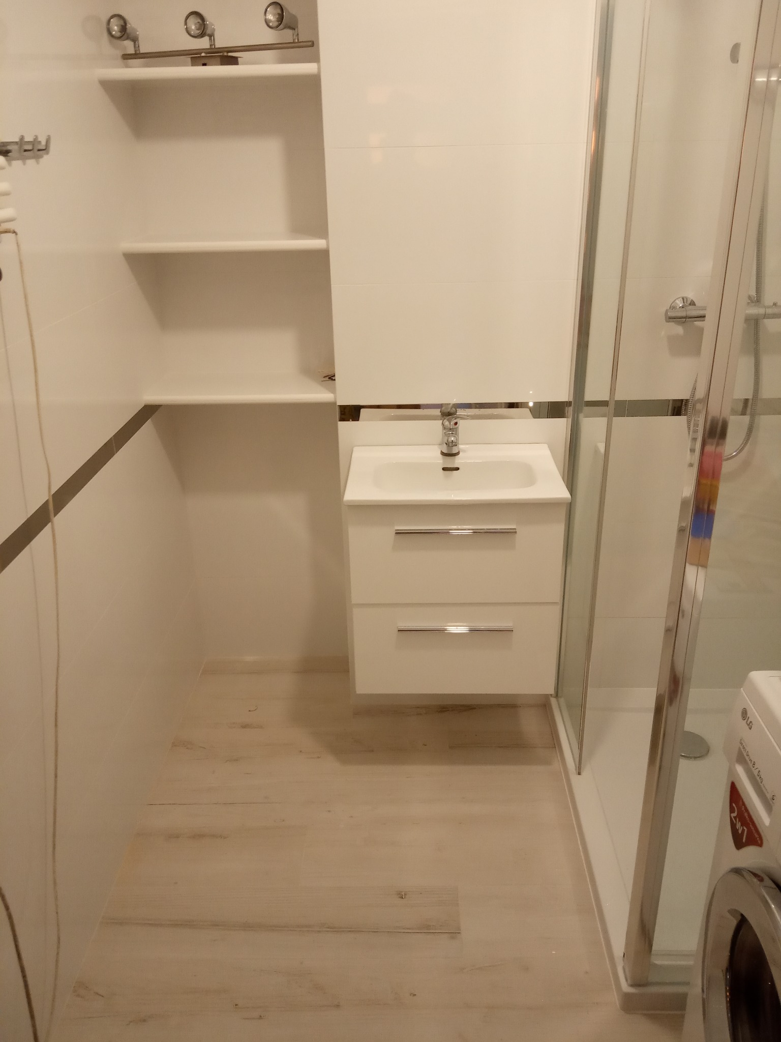 Nowoczesna, jasna łazienka z białą umywalką z szafką, kabiną prysznicową i pralką LG, wykończona płytkami z delikatnym szarym paskiem dekoracyjnym.