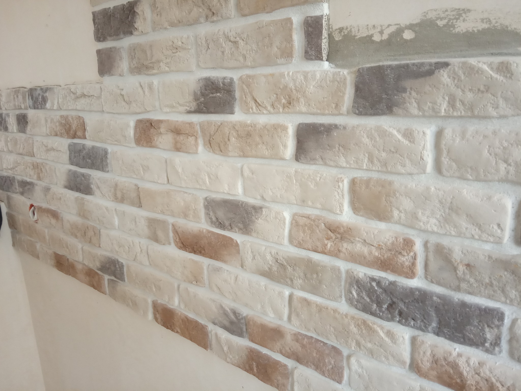 Ściana wyłożona dekoracyjną cegłą w odcieniach bieli, beżu i szarości, z widocznymi spoinami, fragment ściany z niedokończonym wykończeniem u góry.