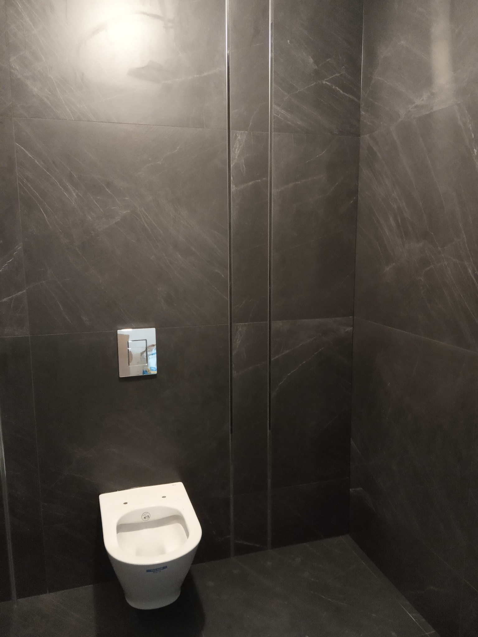 Nowoczesna toaleta podwieszana z chromowanym przyciskiem spłukującym, obłożona ciemnoszarą płytką imitującą marmur z delikatnym żyłowaniem.