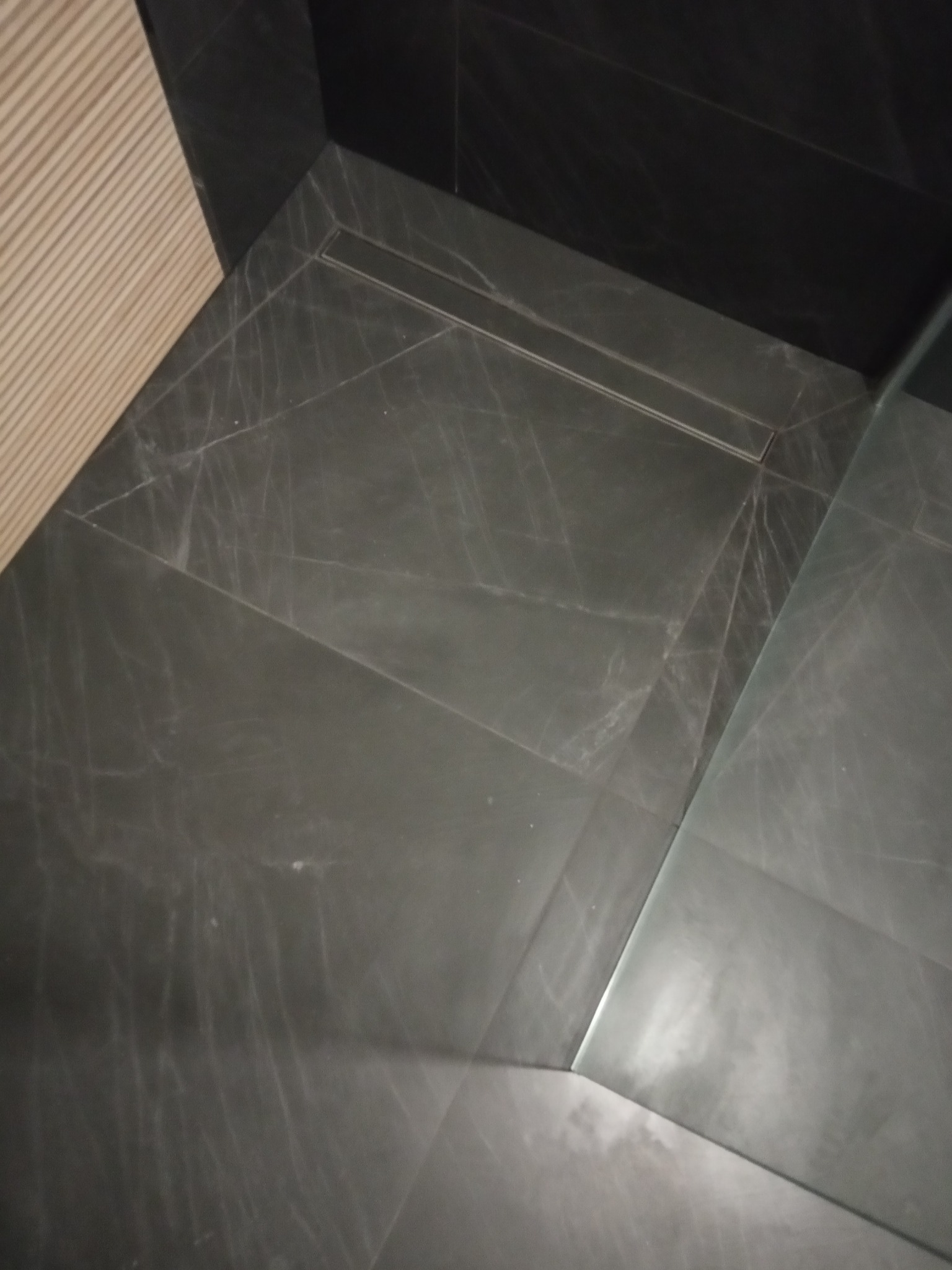 Ciemnoszara, marmurkowa podłoga łazienki z odpływem liniowym i szklaną ścianką kabiny prysznicowej, widok z góry