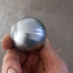 Polerowana, metalowa kula z widocznym wgłębieniem, trzymana w dłoniach na tle betonowej posadzki.