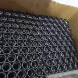 Partie metalowych tulejek z gwintem wewnętrznym, ułożonych równo w kartonowym pudle, widoczne ślady pakowania i transportu.