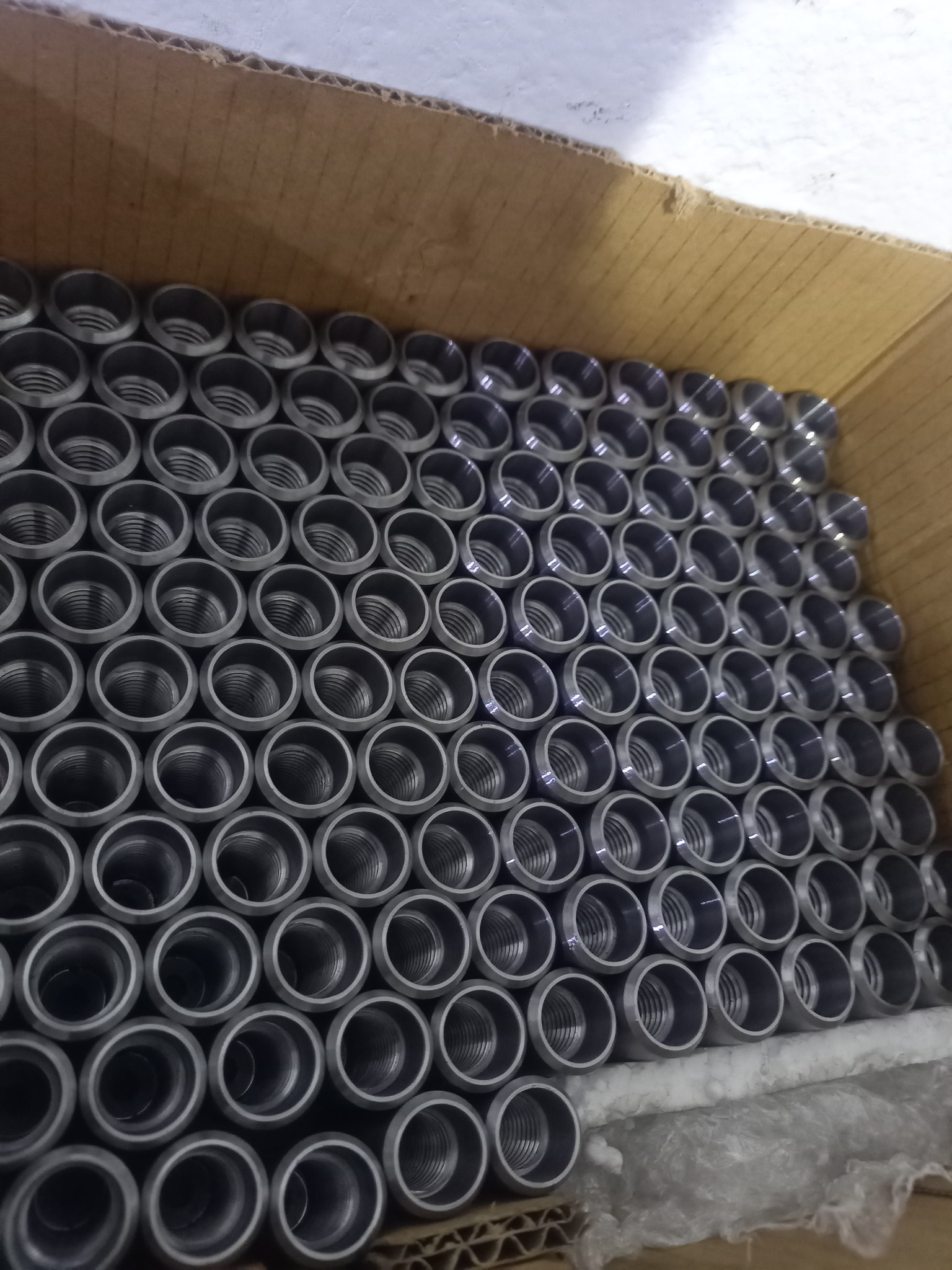 Partie metalowych tulejek z gwintem wewnętrznym, ułożonych równo w kartonowym pudle, widoczne ślady pakowania i transportu.
