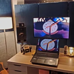 Wnętrze pomieszczenia z panelami akustycznymi, monitory studyjne, laptop z grafiką zegara, gitara akustyczna i mikrofony na biurku.