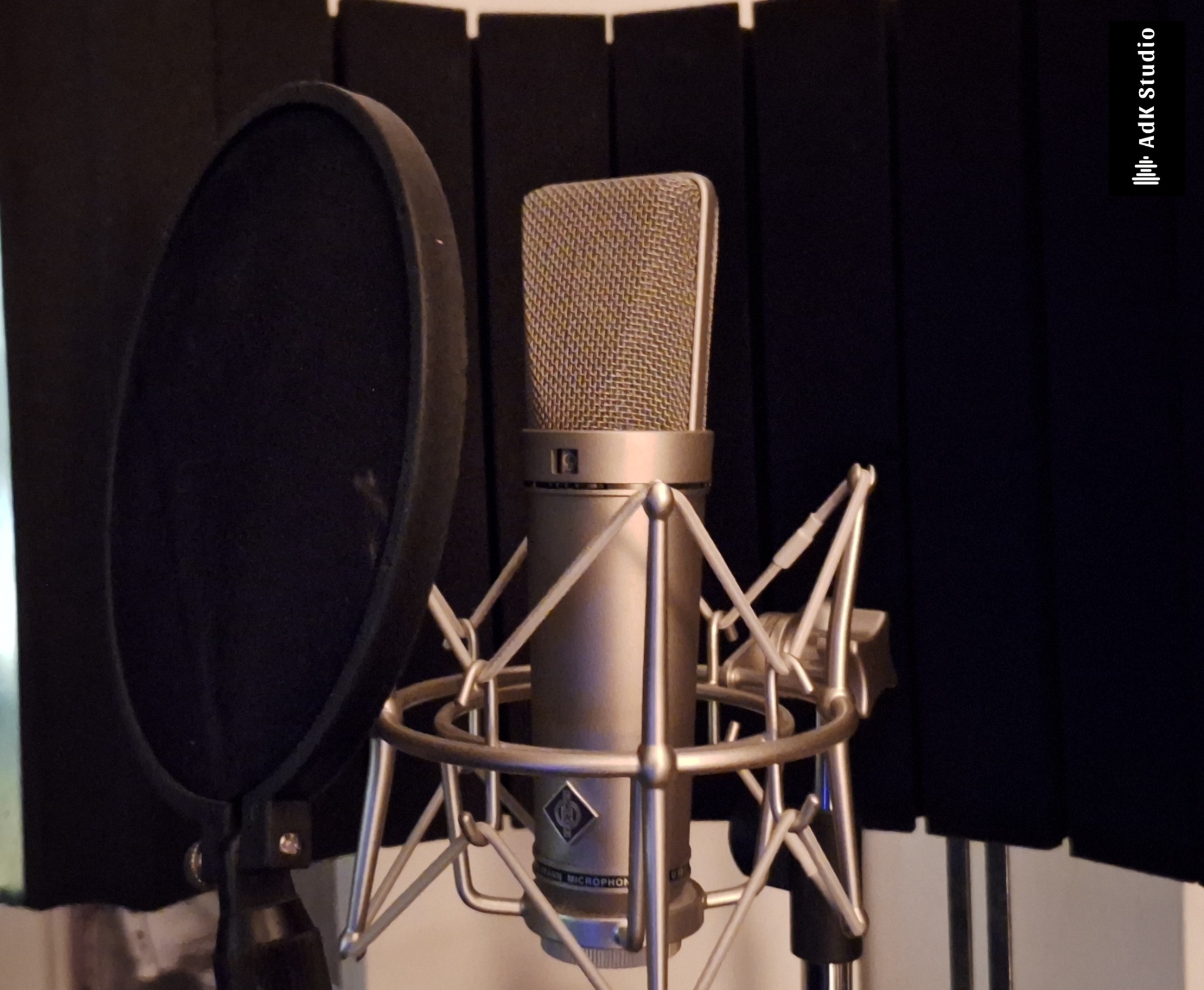 Mikrofon studyjny Neumann z pop filtrem na tle czarnych paneli akustycznych, widoczne logo AdK Studio w prawym górnym rogu.
