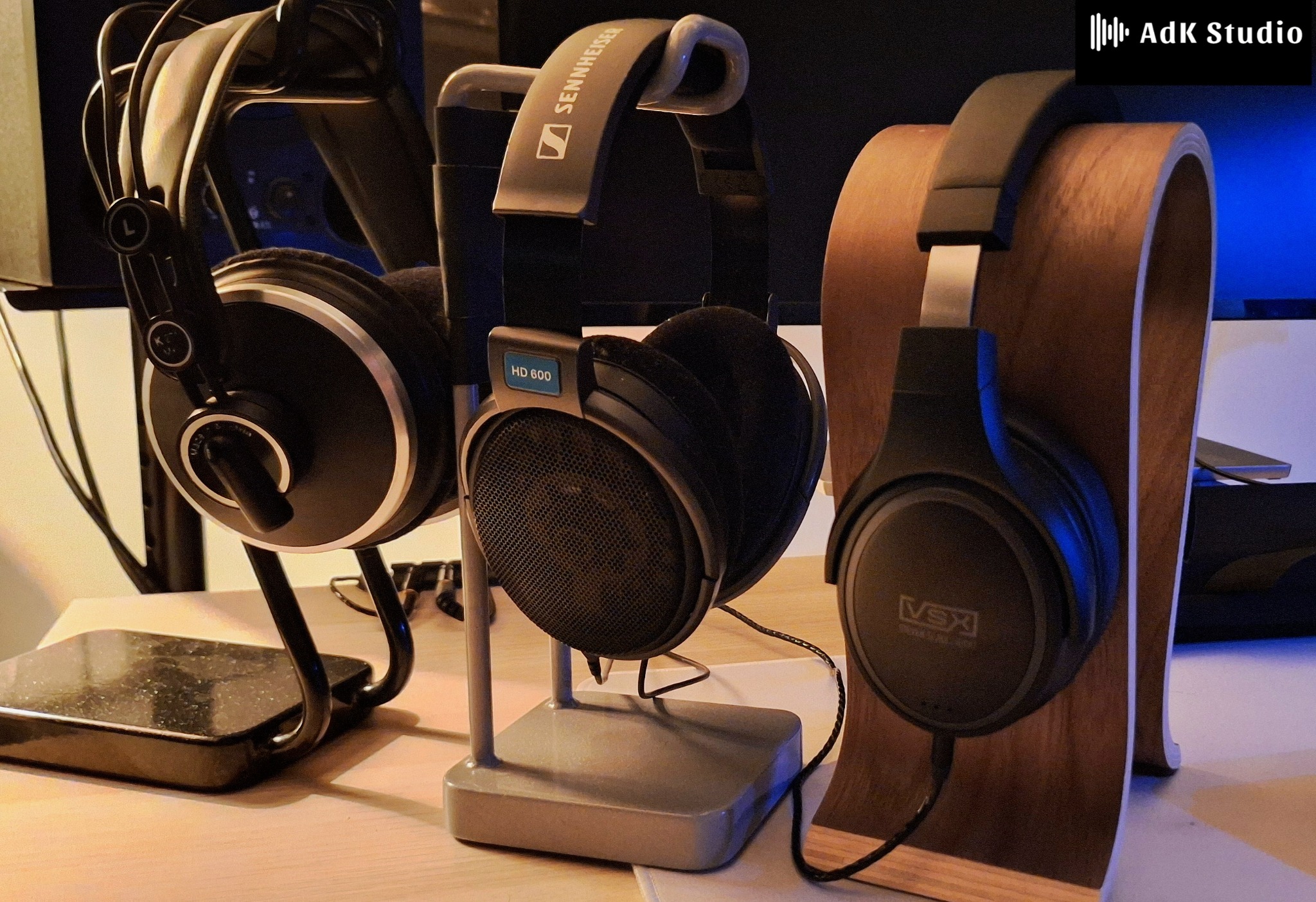 Trzy pary słuchawek studyjnych (AKG, Sennheiser HD 600, VSX) na drewnianych i metalowych stojakach, ustawione na biurku w pomieszczeniu z oświetleniem LED.