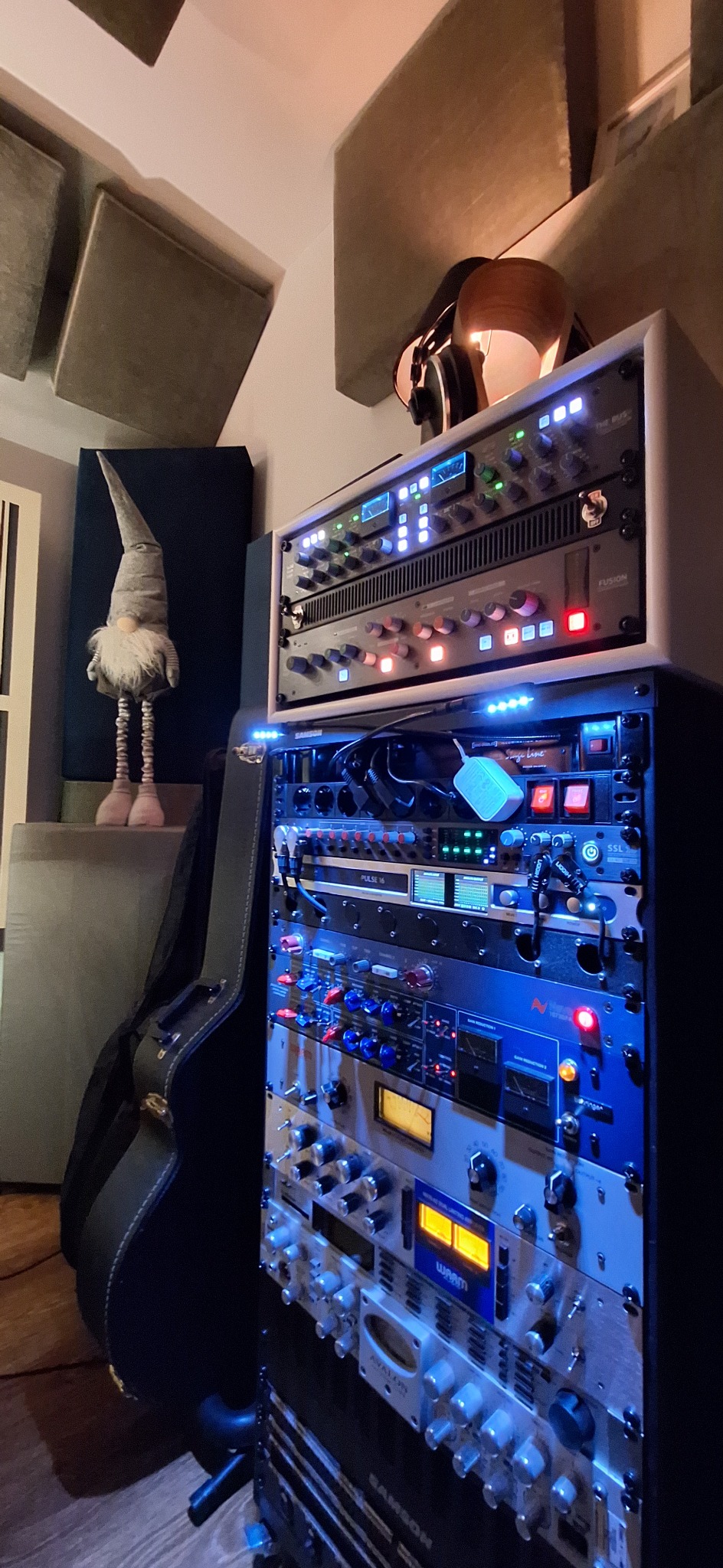 Sprzęt studyjny z podświetleniem LED, obudowa gitary i figurka skrzata w tle. Elementy akustyczne na ścianie. Widok na urządzenia rack w profesjonalnym studio nagrań.