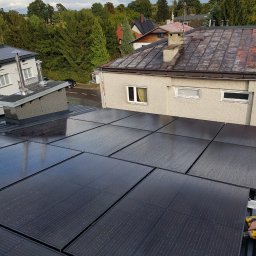 Usługi budowlane - Montaż paneli słonecznych na dachu budynku, widoczne kropelki wody na panelach, w tle zabudowa jednorodzinna i zieleń.