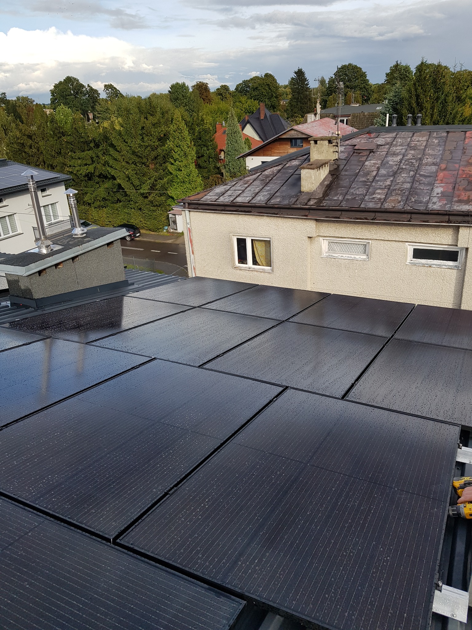 Montaż paneli słonecznych na dachu budynku, widoczne kropelki wody na panelach, w tle zabudowa jednorodzinna i zieleń.