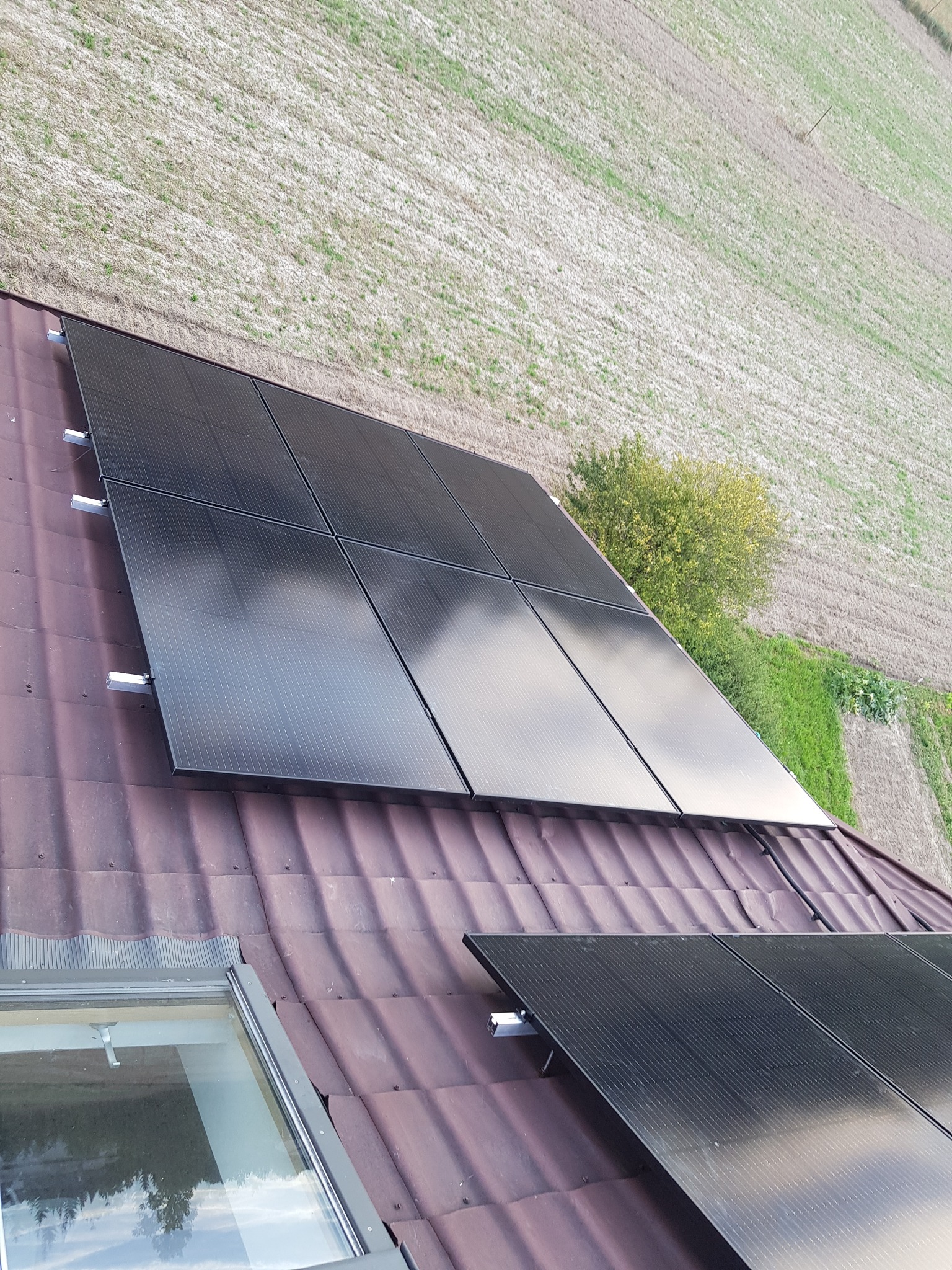 Panele słoneczne zamontowane na dachu z dachówką ceramiczną, widok z góry na pole i krzewy.