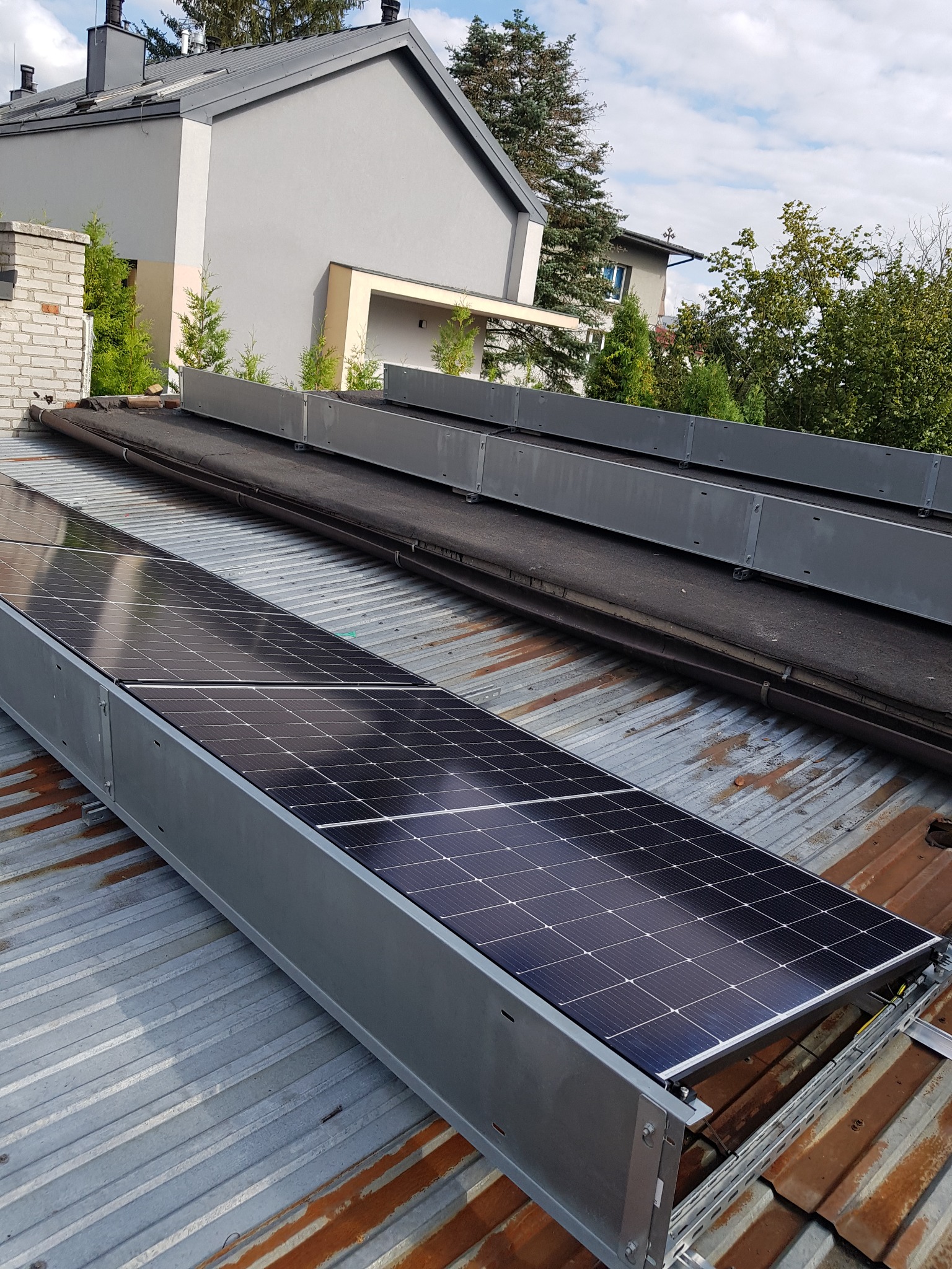 Panele fotowoltaiczne zamontowane na skorodowanej, blaszanej powierzchni dachu, z widocznymi elementami montażowymi i konstrukcją wsporczą.