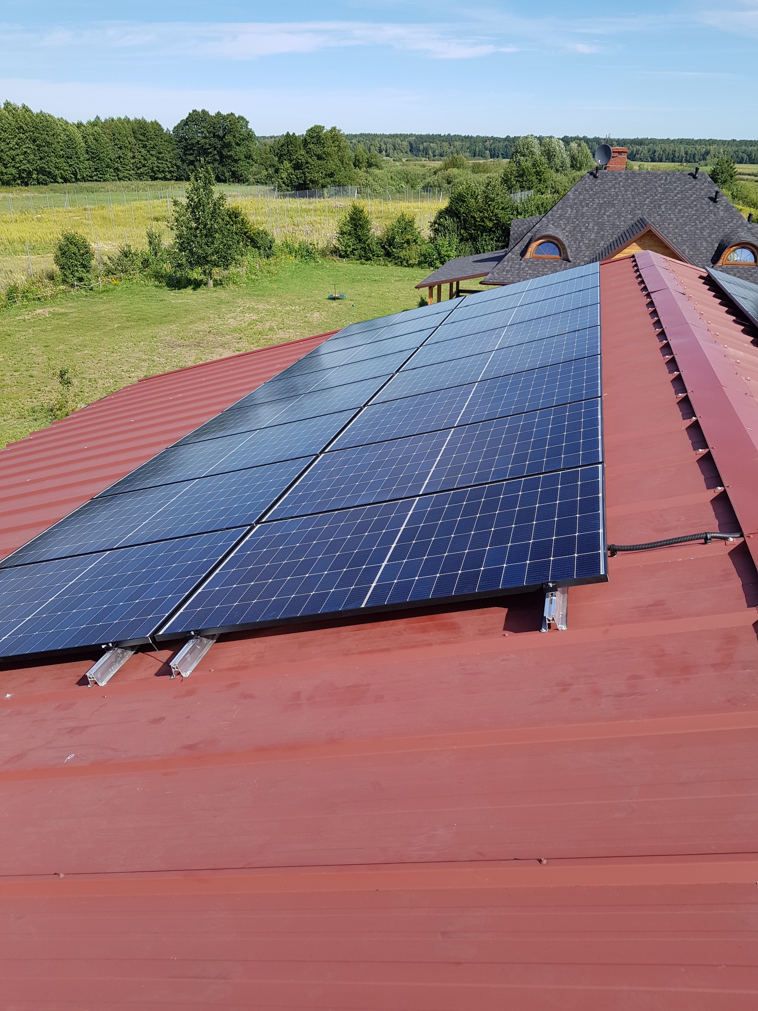 Panele słoneczne zamontowane na czerwonym, metalowym dachu domu jednorodzinnego z widokiem na zielone pola i las w tle.