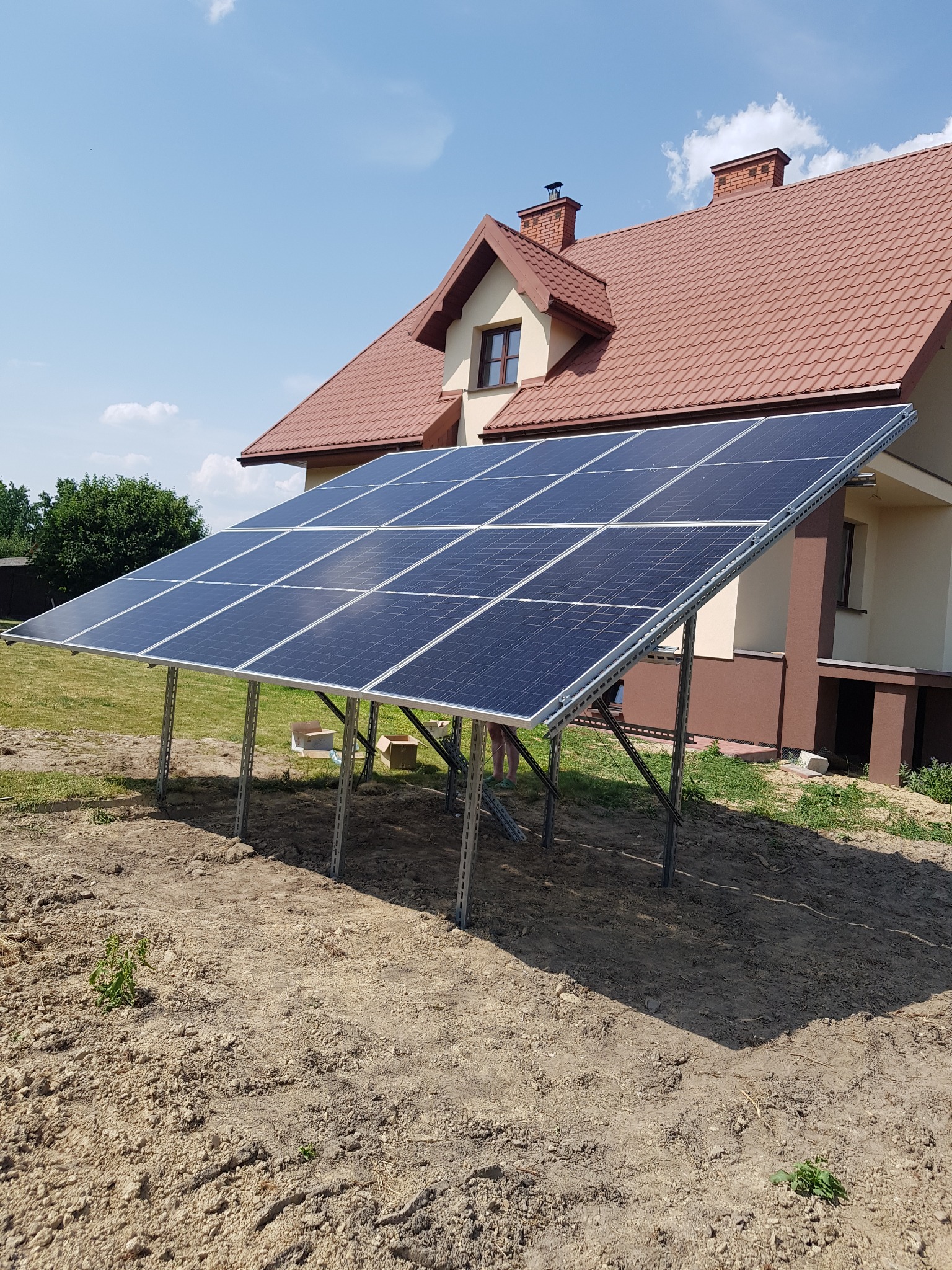 Panele słoneczne na metalowej konstrukcji, ustawione na ziemi przed domem z czerwoną dachówką, w słoneczny dzień.
