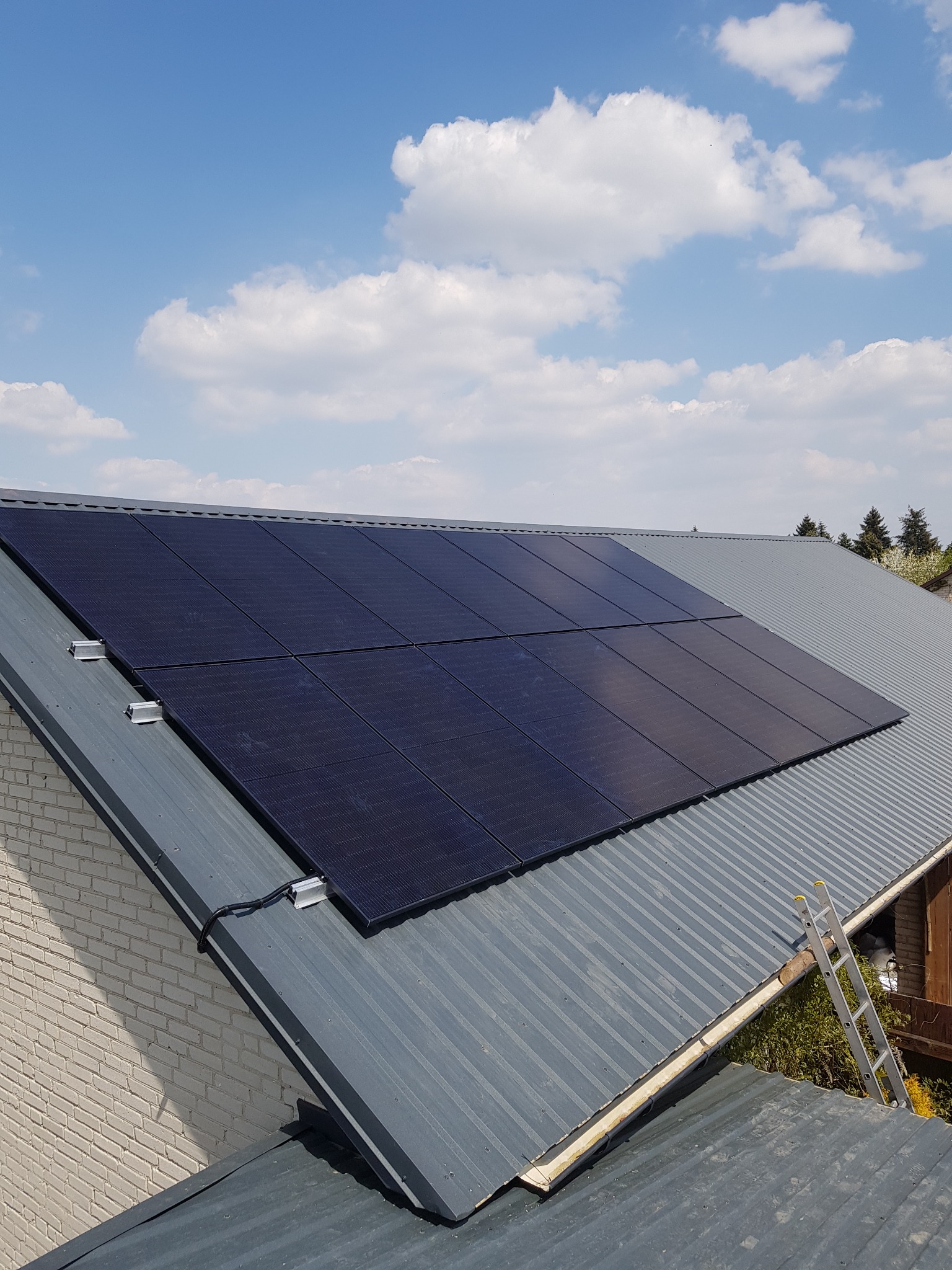 Panele słoneczne zamontowane na szarym, blaszanym dachu z widoczną drabiną opartą o rynnę, na tle błękitnego nieba z białymi chmurami.