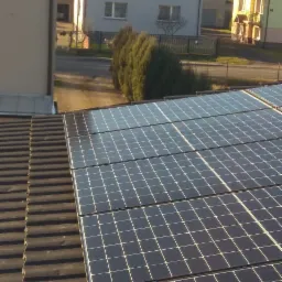 Panele słoneczne na dachu domu jednorodzinnego, widok z góry, w tle budynki mieszkalne i drzewa.