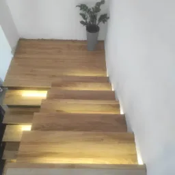 Drewniane schody z podświetleniem LED, widok z góry, z rośliną doniczkową w szarej osłonce przy ścianie, minimalistyczny design.