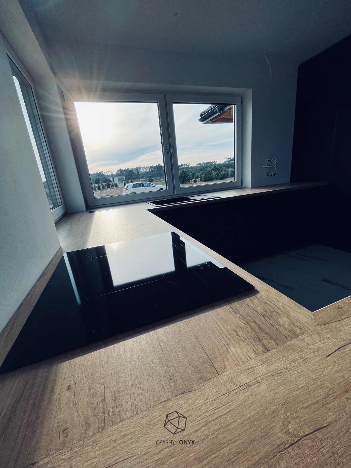 Nowoczesna kuchnia z płytą indukcyjną w jasnym drewnie, widok przez okno na podwórze z białym samochodem, logo firmy w rogu