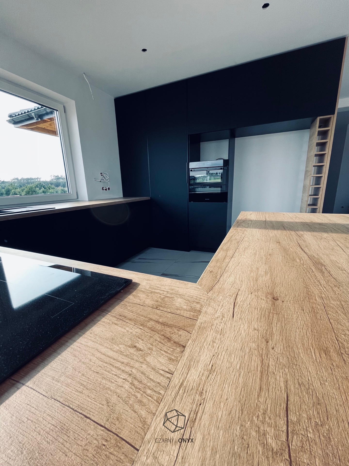 Nowoczesna kuchnia z czarnymi szafkami i blatem z jasnego drewna, widok na okno z zielenią w tle, fragment płyty indukcyjnej i logo firmy w rogu.