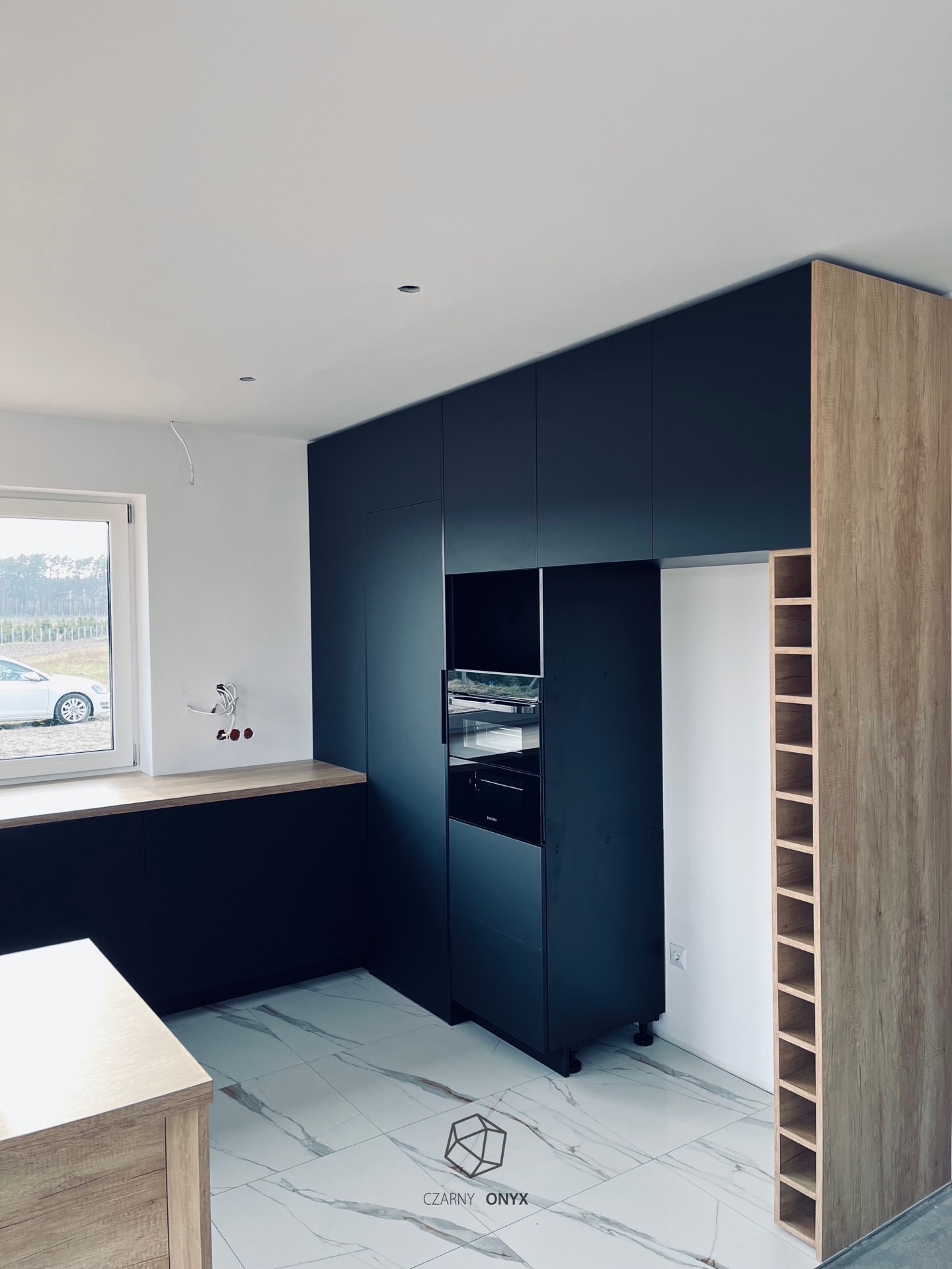 Ciemna zabudowa kuchenna z piekarnikiem i regałem na wino w surowym wnętrzu, widoczne niezabezpieczone kable elektryczne przy oknie.