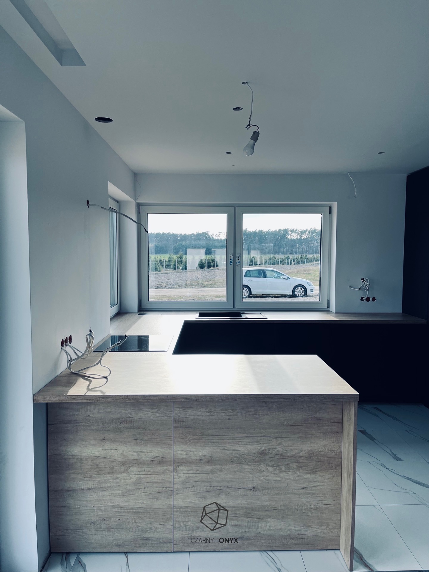 Wyspa kuchenna z drewnianym wykończeniem i czarnym blatem, widok na okno z samochodem na zewnątrz, instalacja elektryczna w trakcie montażu.