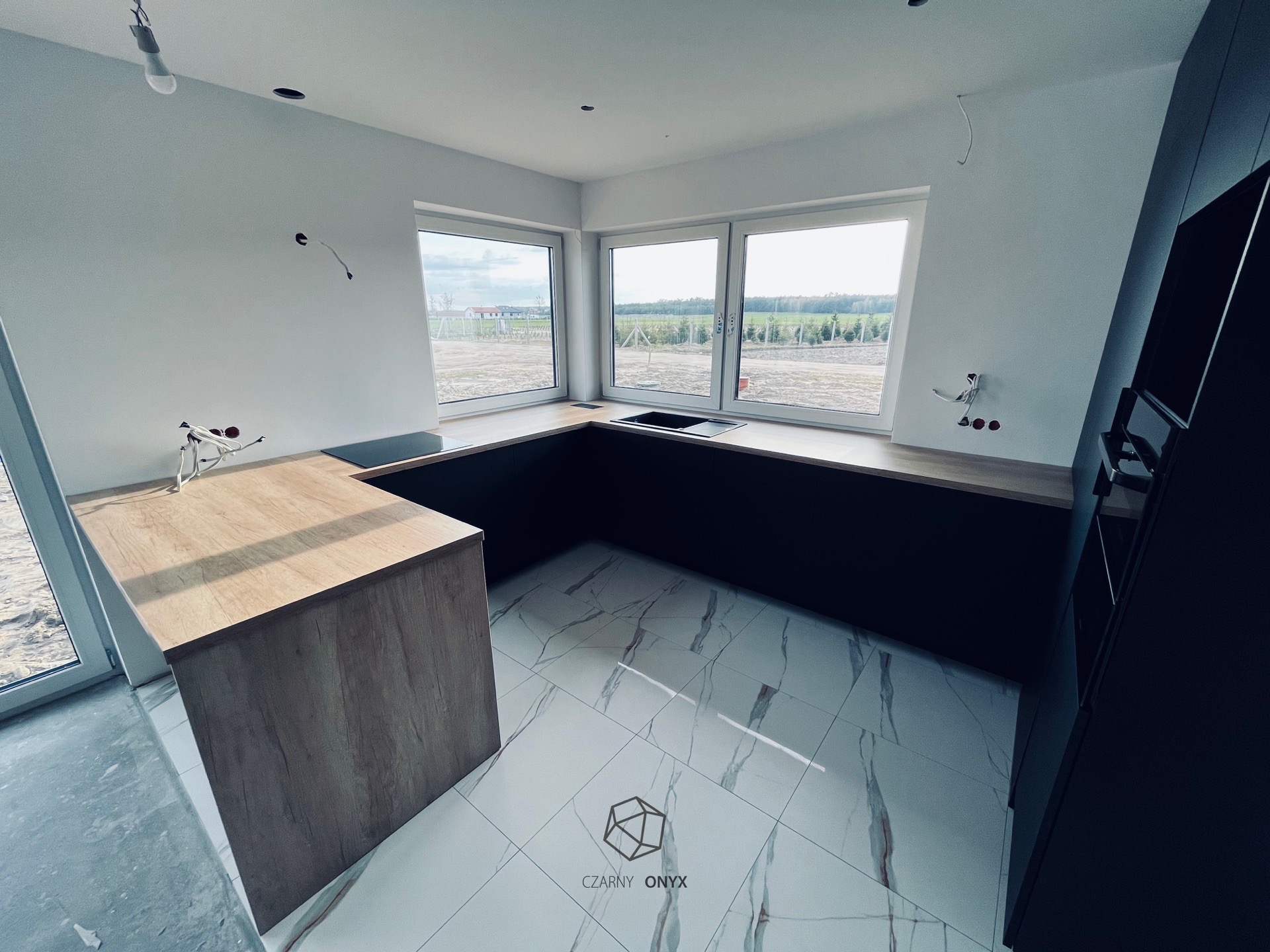 Wnętrze kuchni w trakcie montażu: widoczne niedokończone instalacje elektryczne, surowe ściany, marmurowe płytki podłogowe z logo firmy, zabudowa kuchenna w odcieniach drewna i grafitu...