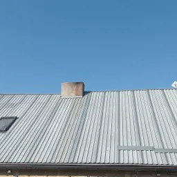 Widok na blaszany dach z kominem, oknem dachowym i anteną na tle błękitnego nieba.