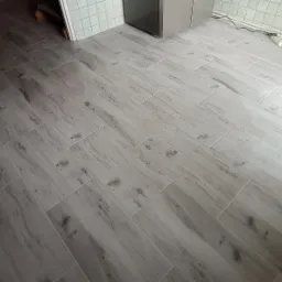 Ułożone pod kątem jasnoszare płytki podłogowe imitujące drewno, widoczny fragment ściany z białymi płytkami, gniazdko elektryczne i butelka wody.