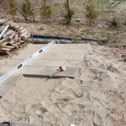 Układanie szarych płyt chodnikowych na piasku, poziomica aluminiowa, gumowy młotek, grabie i stos drewna w tle.