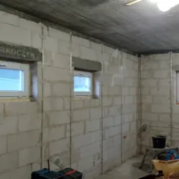 Surowy stan wnętrza w trakcie remontu: ściany z betonu komórkowego z osadzonymi oknami, widoczne instalacje elektryczne, metalowe profile pionowe, betonowy sufit, narzędzia na podłodze.