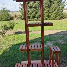 Drewniany, trzypoziomowy kwietnik ogrodowy z dodatkowym małym stołkiem, świeżo pomalowany bejcą, ustawiony na kartonie na trawie, w tle drzewa iglaste i pole.