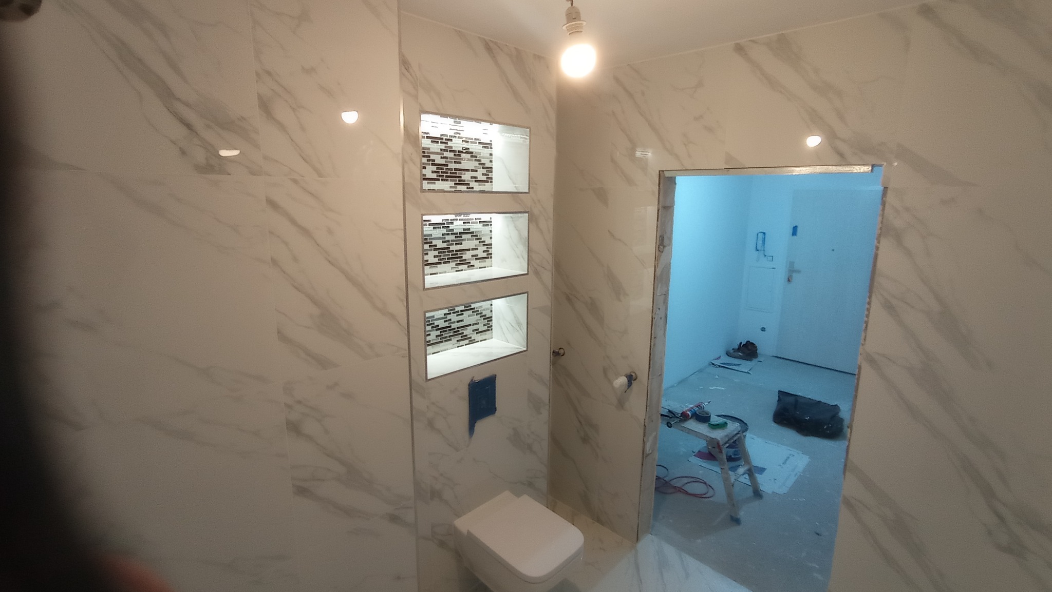 Wykończona łazienka z marmurowymi płytkami, wnękami z mozaiką i podwieszaną toaletą; widoczny fragment pomieszczenia w trakcie prac wykończeniowych przez otwarte drzwi.
