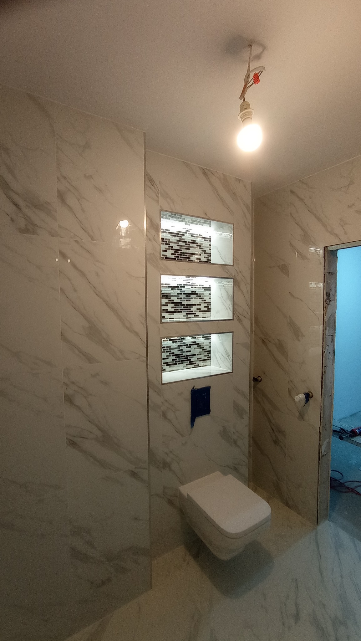 Wykończona łazienka z marmurowymi płytkami, wnękami z mozaiką, podwieszaną toaletą i wiszącą żarówką bez klosza.