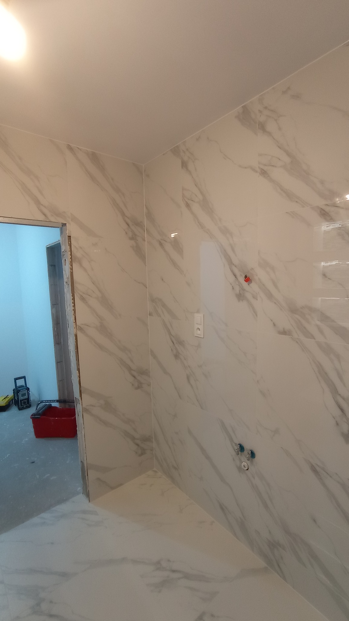 Świeżo wykafelkowana łazienka z płytkami imitującymi marmur, widoczne otwory na baterię i niezamaskowane gniazdko elektryczne, w tle wiadro i radio budowlane.