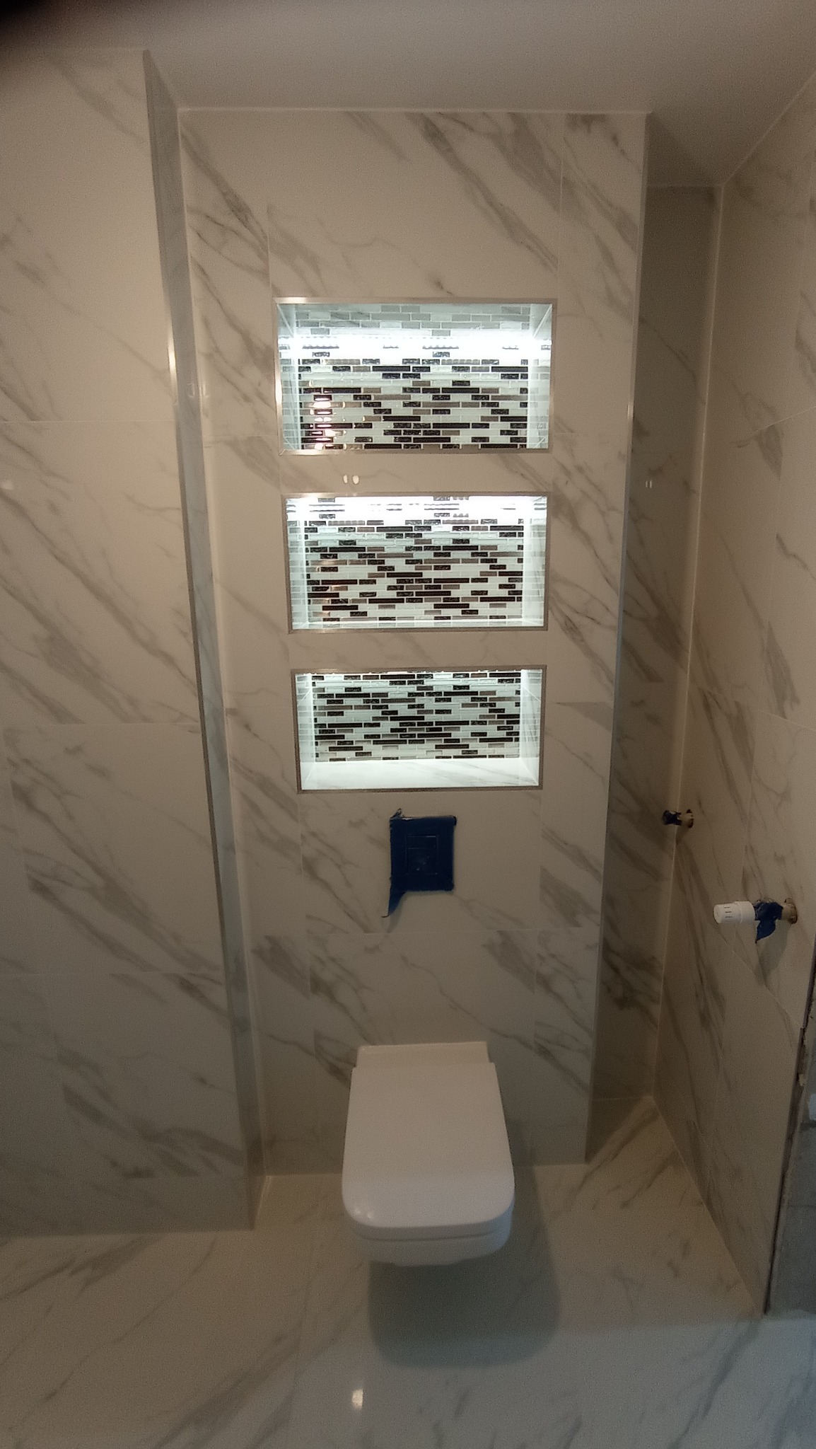 Wykończona łazienka z podwieszaną toaletą i trzema podświetlonymi wnękami z mozaiką na ścianie w marmurowym wzorze.