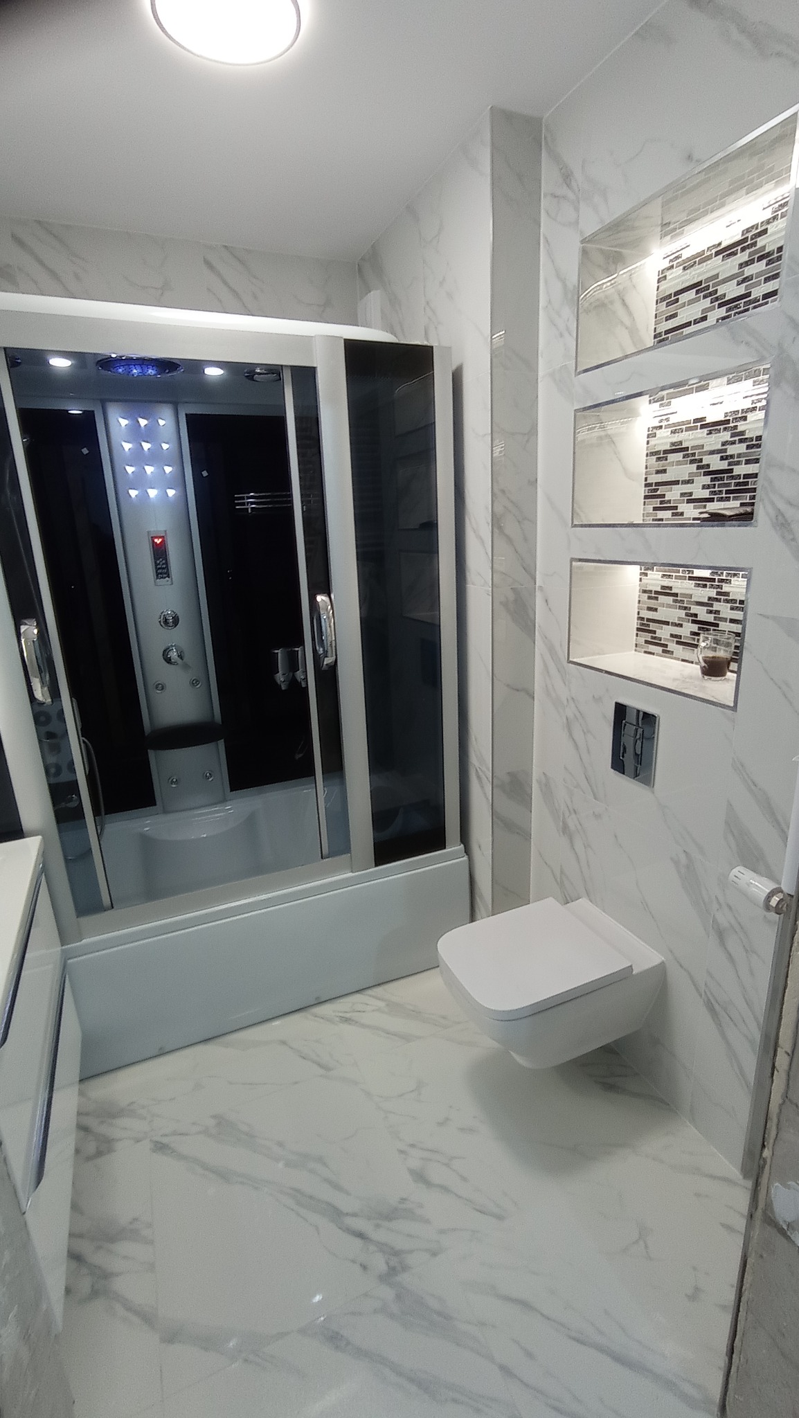 Biała łazienka z kabiną prysznicową z hydromasażem, podwieszaną toaletą i wnękami dekoracyjnymi wyłożonymi mozaiką w odcieniach szarości i bieli, marmurowe płytki na ścianach i podłodze.