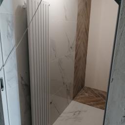 FirmBud - Nowoczesna łazienka z białymi płytkami imitującymi marmur, grzejnikiem drabinkowym i akcentem drewnopodobnych płytek ułożonych w jodełkę na ścianie i podłodze.