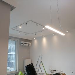 FirmBud - Wnętrze w trakcie remontu z widoczną instalacją oświetleniową na suficie, drabina, zakryty klimatyzator i przygotowane gniazdka elektryczne w ścianie.