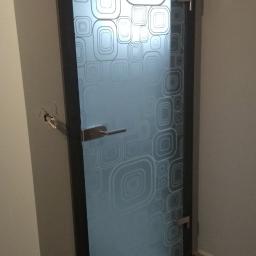 FirmBud - Szklane drzwi z geometrycznym, matowym wzorem kwadratów, zamontowane w ciemnej ramie, z widocznym metalowym uchwytem i zawiasami, osadzone w ścianie z widocznymi śladami po wcześniejszym montażu...