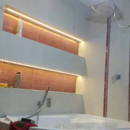 Nowoczesna łazienka z wanną z hydromasażem, podświetlanymi niszami w ścianie wyłożonej płytkami w odcieniach beżu i brązu, oraz szklanym panelem prysznicowym.