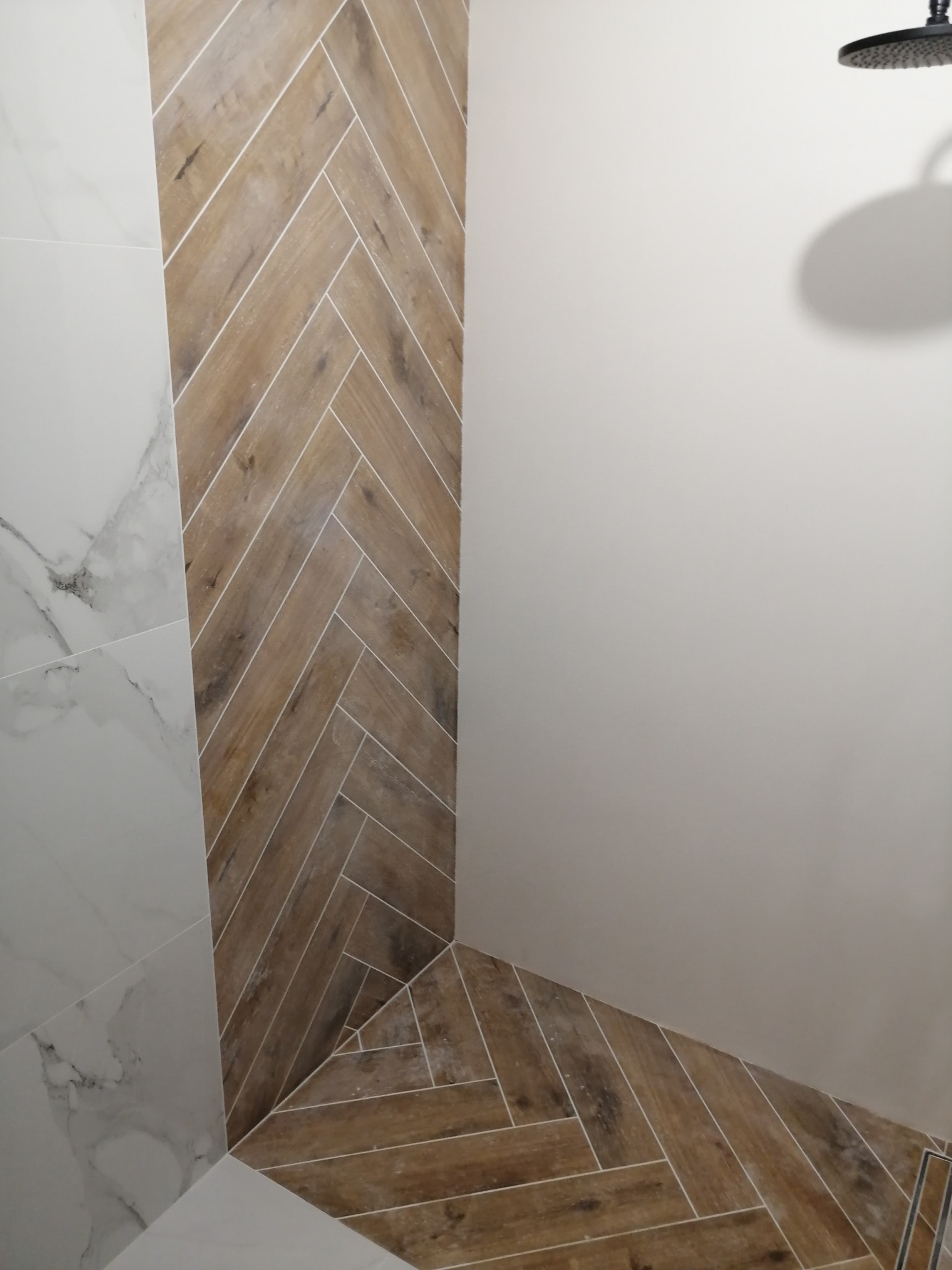 Fragment wykończonej łazienki z drewnopodobnymi płytkami ułożonymi w jodełkę na ścianie i podłodze, kontrastującymi z białymi płytkami imitującymi marmur na ścianie obok oraz widocznym fragmentem...