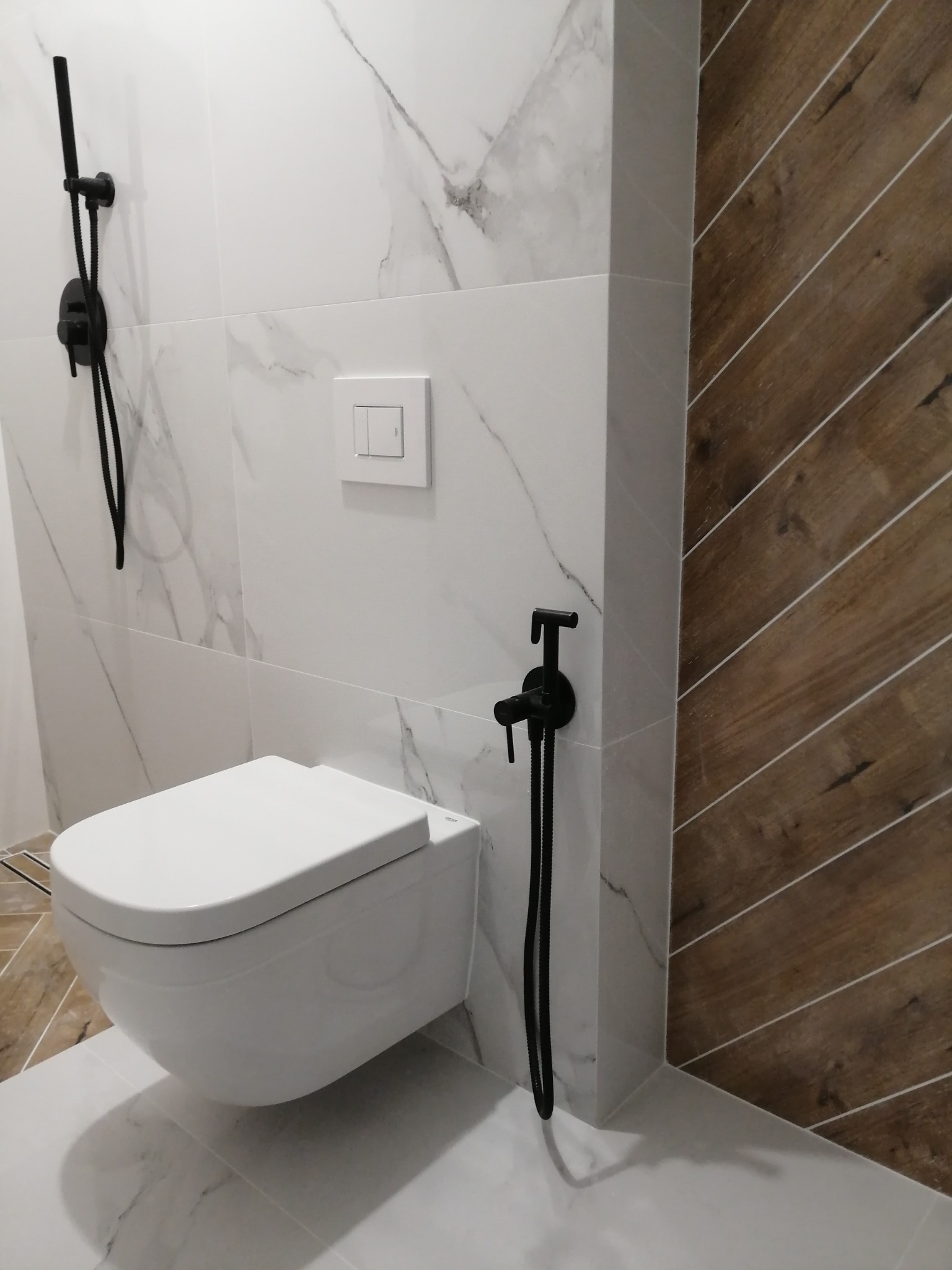 Nowoczesna łazienka z podwieszaną toaletą, białe płytki imitujące marmur i drewnopodobne panele ułożone w jodełkę, czarna armatura.