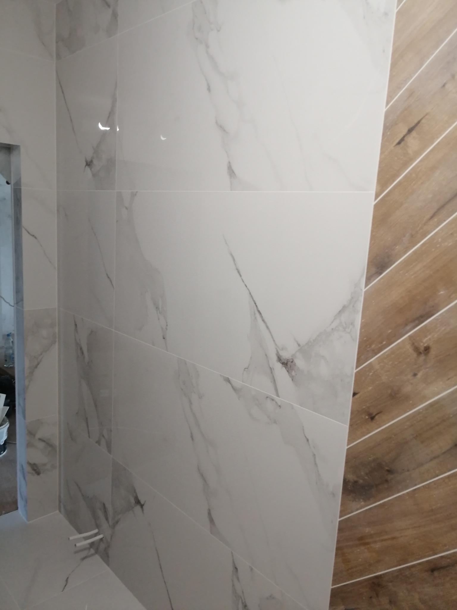 Ściana wyłożona białymi płytkami imitującymi marmur, sąsiadująca z fragmentem ściany wykończonej panelami drewnopodobnymi ułożonymi w jodełkę, widoczne rury wystające ze ściany.