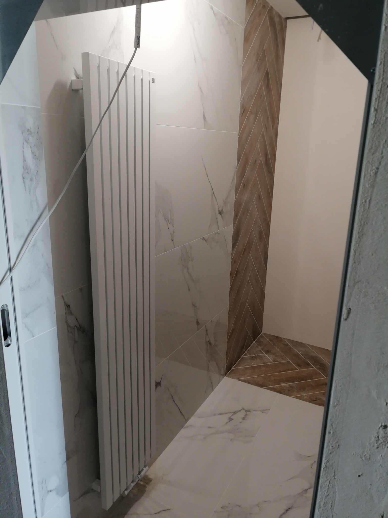 Nowoczesna łazienka z białymi płytkami imitującymi marmur, grzejnikiem drabinkowym i akcentem drewnopodobnych płytek ułożonych w jodełkę na ścianie i podłodze.