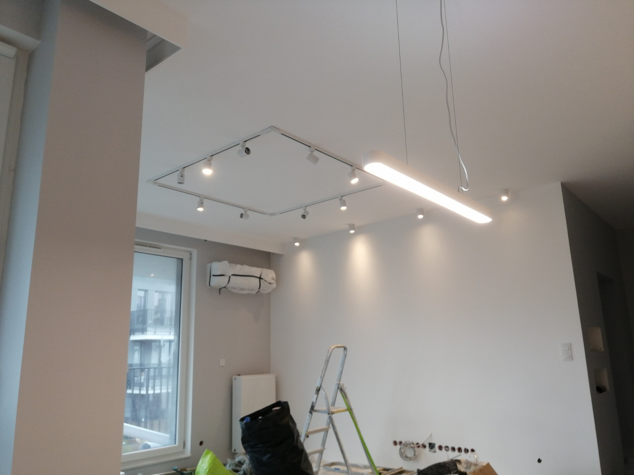 Wnętrze w trakcie remontu z widoczną instalacją oświetleniową na suficie, drabina, zakryty klimatyzator i przygotowane gniazdka elektryczne w ścianie.