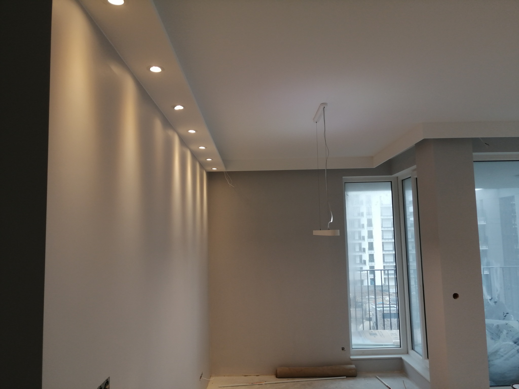 Pomalowane na szaro wnętrze z oświetleniem punktowym w suficie podwieszanym i minimalistyczną lampą wiszącą, widok na osiedle przez duże okno balkonowe.