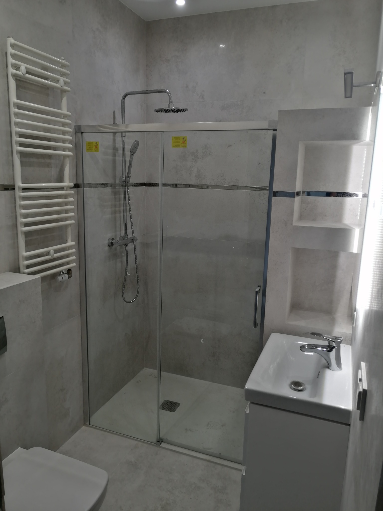 Nowoczesna łazienka w odcieniach szarości z kabiną prysznicową typu walk-in, grzejnikiem drabinkowym i umywalką z szafką, wykończona płytkami imitującymi beton.