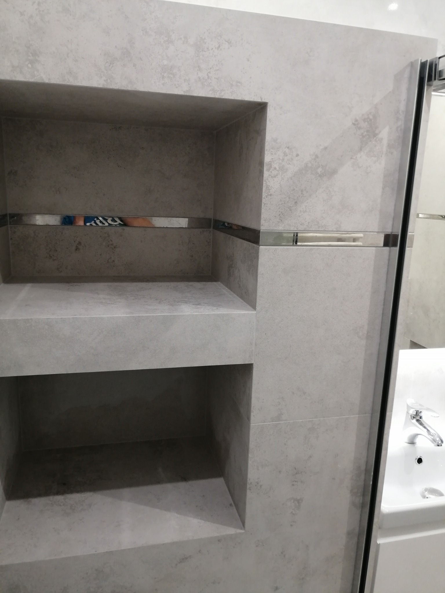 Półki ścienne wyłożone szarą płytką imitującą kamień, z metalowymi listwami dekoracyjnymi, w łazience z białą umywalką i kabiną prysznicową.