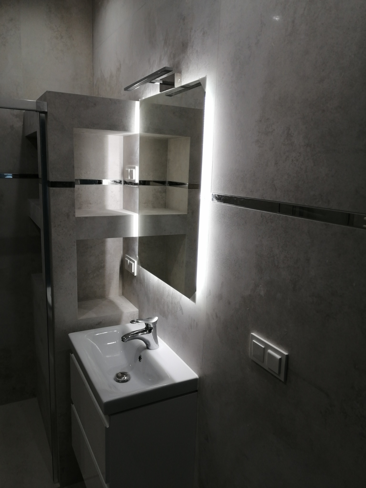 Nowoczesna łazienka z umywalką na szafce, podświetlanym lustrem z półkami wnękowymi i szarą, teksturowaną ścianą z listwami dekoracyjnymi.